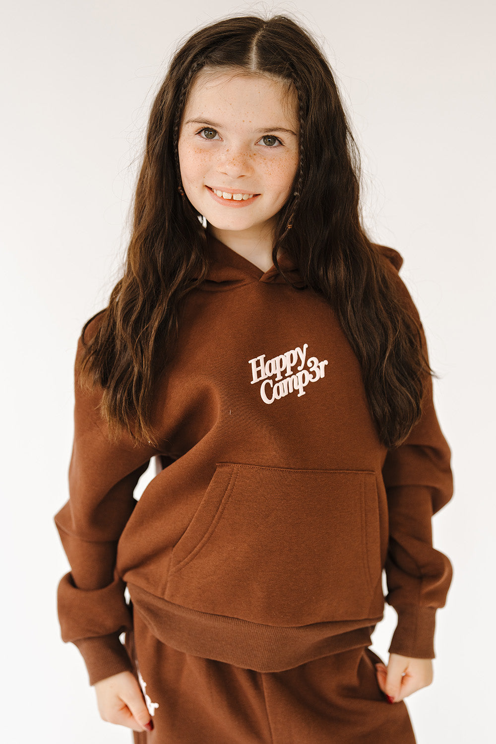 Girl in the espresso Puff Series Core Youth Hoodie with pink chest logo, front view. The Happy Camp3r